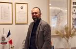 Person med grå kavaj står med glasögon framför en bänk. 